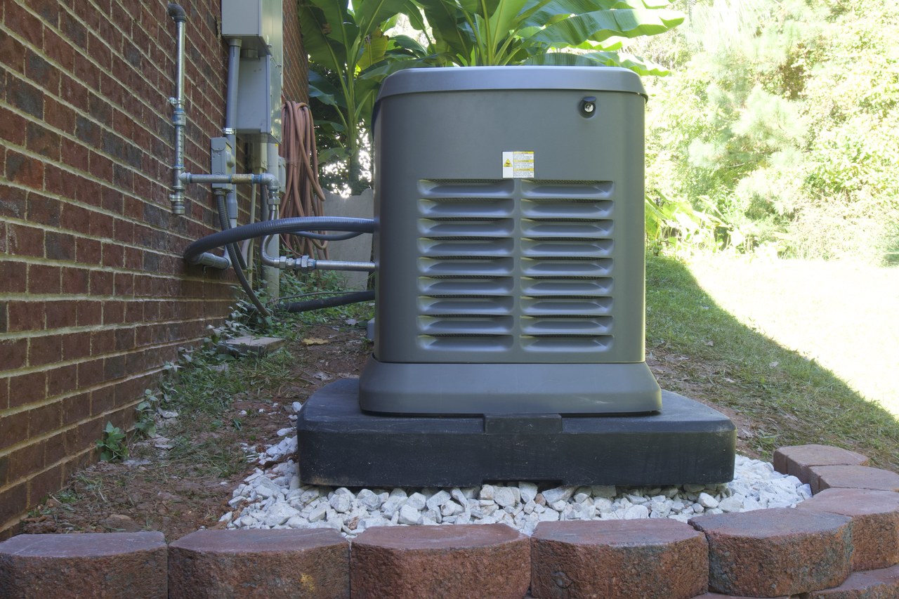Installed home standby generator on a raised pad beside a brick house, with its fuel line and wiring running to wall-mounted electrical equipment on the house